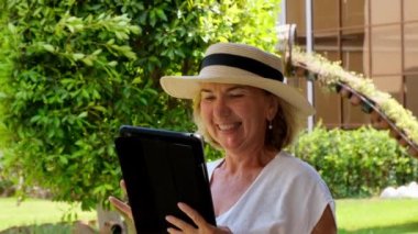 Şapkalı kıdemli sarışın iş kadını, iş için tablet, dijital tablet ya da kendi yeşil bahçesinde eğitim görüyor. 50-55 yaşlarında bir kadın. Online iş ve eğitim kavramları