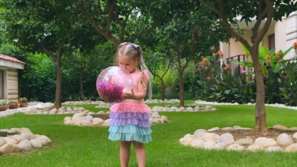 Милая маленькая девочка, 3 года, с двумя хвостиками на голове, одета в тонкое и разноцветное платье розового голубого цвета, играет с ярким прозрачным шаром с разноцветными перьями внутри — стоковое видео