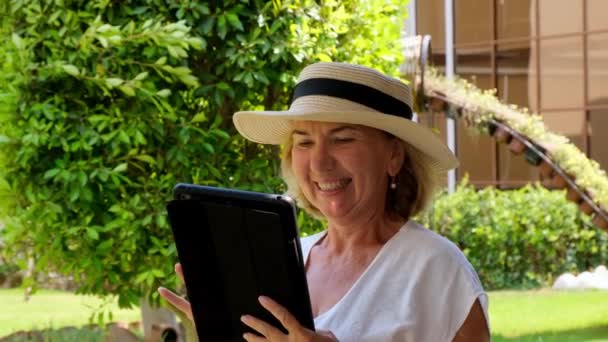 Senior mujer rubia mujer de negocios en sombrero utiliza tableta PC, tableta digital para el trabajo de negocios o estudio en su propio jardín verde. mujer de 50 a 55 años. El concepto de su propio negocio y formación, en línea — Vídeo de stock