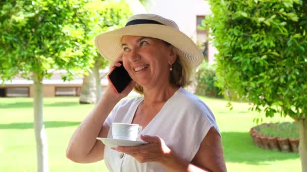 Zabawna blondynka, w wieku 50-55 lat, w kapeluszu i białej sukience, rozmawia za pomocą smartfona i trzyma filiżankę kawy w dłoniach na tarasie w słoneczny poranek w letni dzień. Wolny strzelec — Wideo stockowe