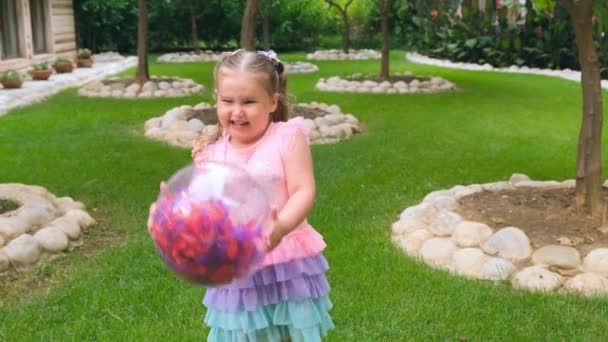Щаслива маленька дівчинка, 3 роки, з двома хвостами на голові, одягнена в ніжну і різнокольорову сукню рожевого синього кольору, грає з яскравим прозорим м'ячем з різнокольоровим пір'ям — стокове відео