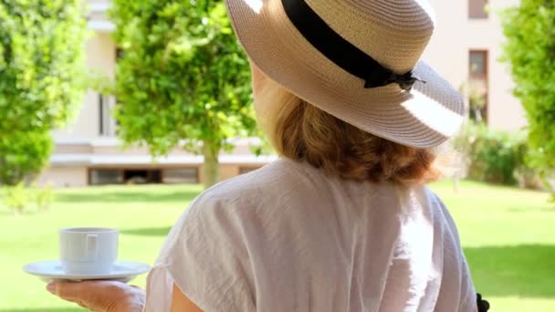 藁帽子の幸せな女性は晴れた日の夏のテラスで朝に座って、熱い飲み物と飲み物のコーヒーのカップを保持しています。お茶を飲むときはマナーがいい。良い朝、旅行の概念 — ストック動画