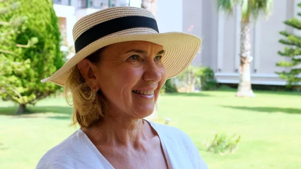 Retrato de uma mulher madura 55-60 anos com um sorriso de dente em um chapéu de palha protetor solar desfrutando de seu tempo contra o pano de fundo de um jardim verde. close-up. — Fotografia de Stock