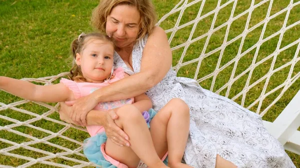 Cheerful Couple grandmother and granddaughter 3 years old are relaxing and basking in a hammock on a green meadow in their garden on a summer sunny day. Vacation concept, generational relationship — Stock Photo, Image
