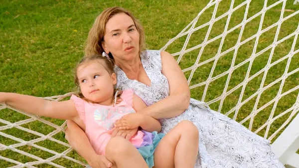 Divertente coppia nonna e nipote 3 anni sono rilassanti e crogiolarsi in un'amaca su un prato verde nel loro giardino in una giornata di sole estivo. Concetto di vacanza, rapporto generazionale — Foto Stock