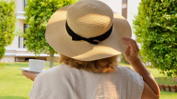Счастливая женщина в соломенной шляпе держит чашку горячего напитка и пьет кофе, сидя утром на летней террасе в солнечный день. Хорошие манеры, когда пьёшь чай. Понятие доброе утро, путешествия — стоковое фото
