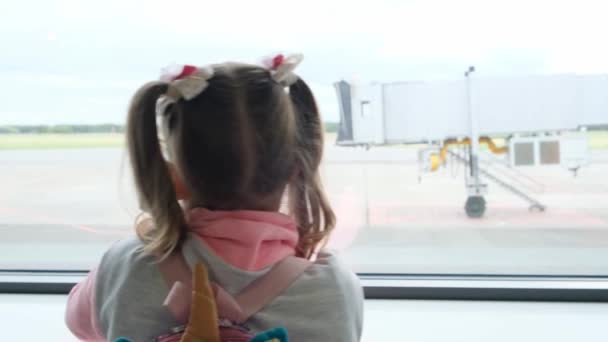 Liten flicka i åldern 5 år tittar ut genom fönstret på flygplatsen, väntar på avgång eller ankomst av planet, drömmer om en resa — Stockvideo