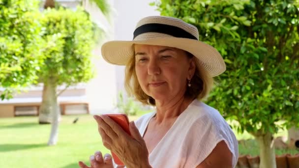 50 세 ~ 55 세 의 금발 세노라 여성 모자와 흰색 드레스를 입고, 스마트폰으로 비디오 통화를 하고, 문자를 입력하고, 맑은 아침 테라스에서 인터넷 정보를 찾는다. — 비디오