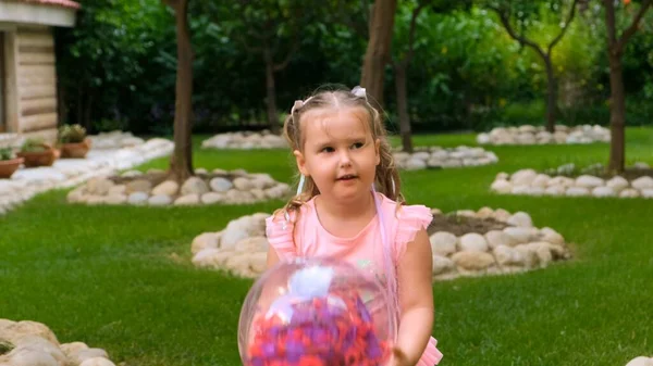 Menina engraçada, 3 anos, com dois rabos-de-cavalo na cabeça, vestida com um vestido delicado e multicolorido de cor azul rosa, joga com uma bola transparente brilhante com penas multicoloridas — Fotografia de Stock