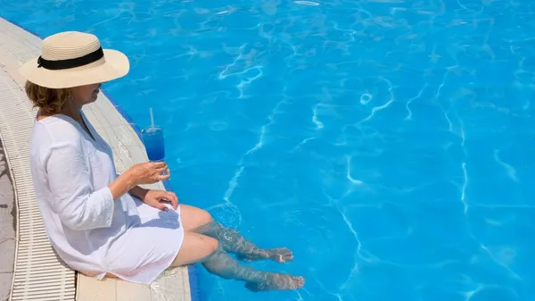 屋外スイミングプールで足をぶら下げている年配の女性をリラックスさせ、麦藁帽子をかぶった青いさわやかなカクテルを持っています。夏休みだ。包括的 — ストック写真