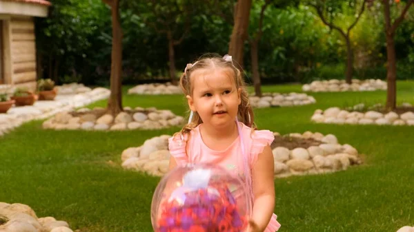 Menina feliz, 3 anos, com dois rabos de cavalo na cabeça, vestida com um vestido delicado e multicolorido de cor azul rosa, joga com uma bola transparente brilhante com penas multicoloridas — Fotografia de Stock