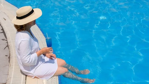青い屋外スイミングプールの近くでリラックスしている幸せなシニア女性青いカクテルを身に着けているわらの帽子。人々は夏休みを楽しんでいます。包括的 — ストック写真