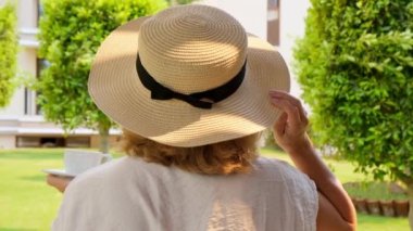 Hasır şapkalı mutlu kadın elinde bir fincan sıcak içecek ve kahve tutuyor, güneşli bir günde yaz terasında oturuyor. Çay içerken görgülü ol. Günaydın, seyahat konsepti.