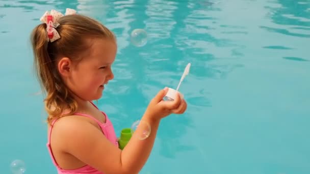 Linda niña de 4-5 años, con colas de caballo, con un traje de baño rosa, soplando bolas de jabón en la piscina en las vacaciones de verano — Vídeos de Stock
