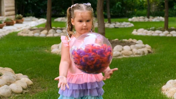 幸せな女の子, 3歳,彼女の頭の上に2つのポニーテールと,ピンクブルーの色の繊細で多色のドレスに身を包んだ,多色の羽を持つ明るい透明ボールで再生されます — ストック写真