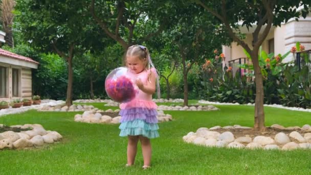 Красива смішна маленька дівчинка, 3 роки, з двома хвостами на голові, одягнена в ніжну і різнокольорову сукню рожевого синього кольору, грає з яскравим прозорим м'ячем з різнокольоровим — стокове відео