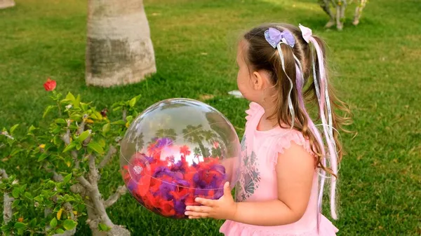 滑稽的小女孩，3岁，头上戴着两条马尾辫，穿着一件淡淡的、多彩的粉色蓝色连衣裙，玩着一个色彩斑斓的透明球，头戴多彩的羽毛 — 图库照片