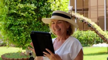 Şapkalı kıdemli sarışın iş kadını yöneticiliği, iş için tablet, dijital tablet veya kendi yeşil bahçesinde eğitim görüyor. 50-55 yaşlarında bir kadın. Kendi iş ve eğitimlerinin konsepti