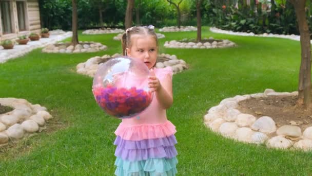 Niña, 3 años de edad, está jugando una pelota inflable transparente con plumas de colores en el interior, con un vestido de arco iris. El concepto de vacaciones de verano, juegos en el hogar, el desarrollo y la educación de — Vídeos de Stock