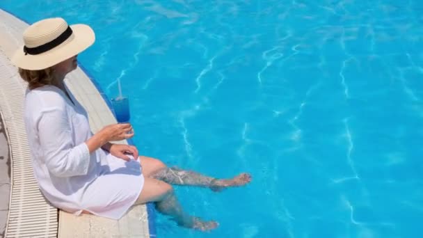 Femme âgée de luxe pendre ses jambes dans une piscine extérieure tenant un cocktail rafraîchissant bleu portant un chapeau de paille. Repos d'été. Tout compris — Video