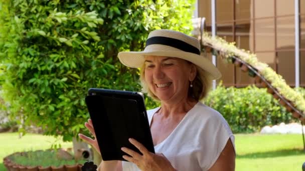 Zarządzanie starszą blondynką bizneswoman w kapeluszu używa tabletu, tabletu cyfrowego do pracy w biznesie lub nauki w swoim zielonym ogrodzie. kobieta w wieku 50-55 lat. Koncepcja własnej działalności gospodarczej i szkolenia — Wideo stockowe