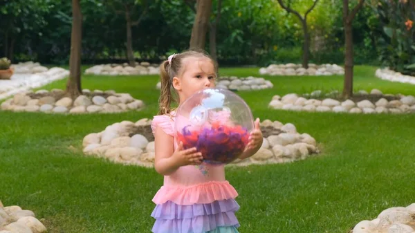 彼女の頭に2本のポニーテールと3歳の陽気な女の子は、ピンクブルーの色の繊細で多色のドレスを着て、多色の羽を持つ明るい透明なボールで遊んでいます — ストック写真