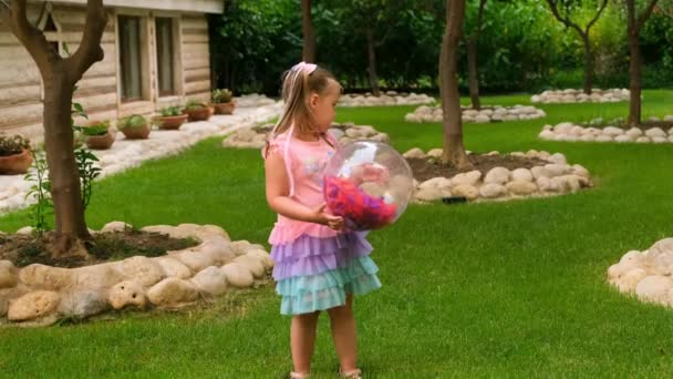 Маленька дівчинка, 3 роки, грає на надувному прозорому м'ячі з барвистим пір'ям всередині, одягнений у веселкове плаття. Концепція літніх канікул, ігор вдома, розвиток та освіта — стокове відео