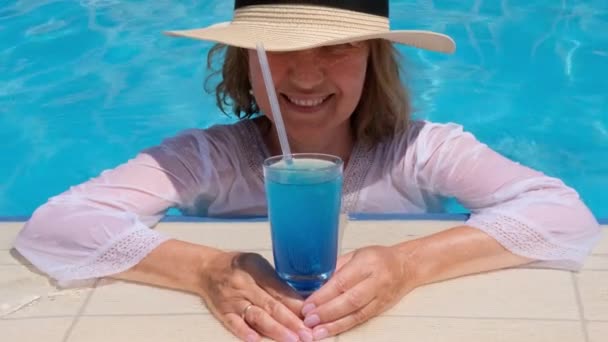 Eine fröhliche Seniorin entspannt sich in der Nähe des blauen Freibades mit blauem Cocktail und Strohhut. Die Menschen genießen ihren Sommerurlaub. All inclusive — Stockvideo