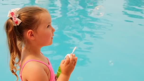 Niedliches kleines Mädchen, 4-5 Jahre alt, mit Pferdeschwanz, trägt einen rosafarbenen Badeanzug, bläst im Sommerurlaub Seifenbälle im Pool — Stockvideo