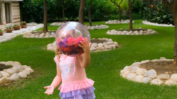 Vrolijk klein meisje, 3 jaar oud, met twee paardenstaarten op haar hoofd, gekleed in een delicate en multi-gekleurde jurk van roze blauwe kleur, speelt met een heldere transparante bal met multi-gekleurde veren — Stockfoto