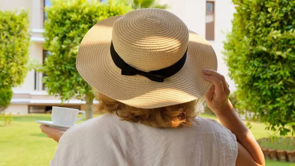 Женщина в возрасте 50-55 лет в соломенной шляпе держит чашку горячего напитка и пьет кофе, сидя утром на летней террасе в солнечный день. Концепция доброго утра, путешествия и ожидания нового — стоковое фото