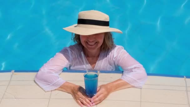 Lächelnde Seniorin entspannt sich in der Nähe des blauen Freibades bei einem blauen Cocktail. Die Menschen genießen ihren Sommerurlaub — Stockvideo