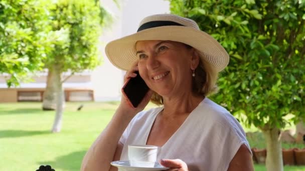 Милая блондинка-сеньора, 50-55 лет, в шляпе и белом платье, разговаривает с помощью смартфона и держит чашку кофе в руках на террасе в солнечное утро в летний день. Фрилансер — стоковое видео