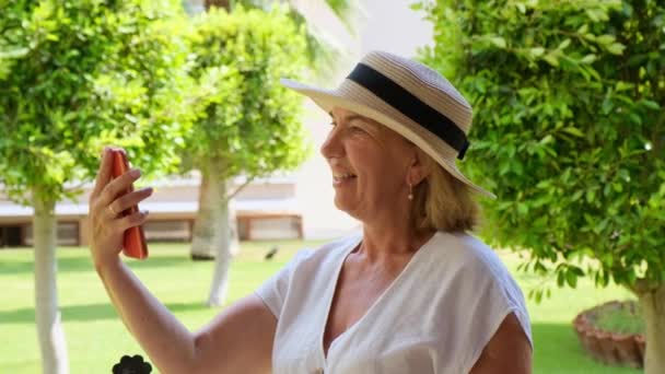 金髪の50 〜 55歳の女性、帽子と白いドレスで、夏の日の晴れた朝に彼女のテラスでビデオ通話でスマートフォンを使用して話をします。フリーランスのコンセプト、女性ブロガー、ビジネス — ストック動画