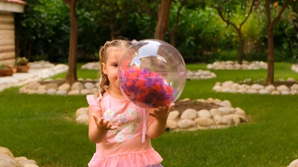 Смешная маленькая девочка, 3 года, с двумя хвостиками на голове, одета в тонкое и разноцветное платье розового голубого цвета, играет с ярким прозрачным шаром с разноцветными перьями — стоковое фото