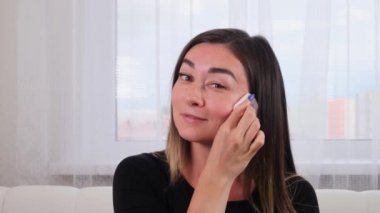 Genç bir kız kusursuz cildini bir süngerle temizler ve aynanın önünde beyaz bir pamuk ped kullanır. Genç bir kadın yüzüne bakar. Spa ve sağlık, cilt bakımı konsepti. Kişisel bakım