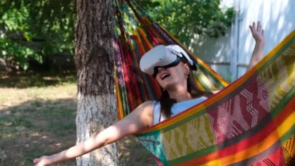 Mujer alegre con gafas de realidad virtual descansa en una hamaca cerca de un árbol, imaginando un sueño hecho realidad, un mundo inventado. El concepto de adquirir experiencia utilizando gafas de realidad virtual — Vídeos de Stock