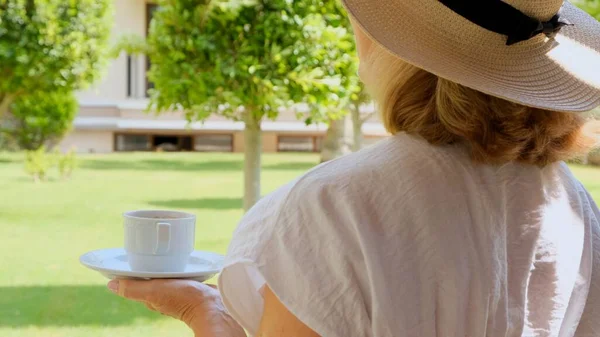 Hasır şapkalı mutlu kadın elinde bir fincan sıcak içecek ve kahve tutuyor, güneşli bir günde yaz terasında oturuyor. Çay içerken görgülü ol. Günaydın, seyahat konsepti. — Stok fotoğraf