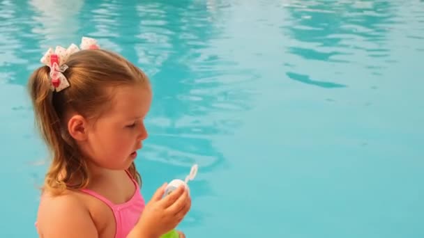 Смешная маленькая девочка 4-5 лет, одетая в розовый купальник, дующая мыльные шарики в бассейн на летних каникулах — стоковое видео