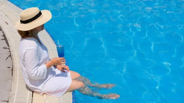 屋外スイミングプールで足をぶら下げている年配の女性をリラックスさせ、麦藁帽子をかぶった青いさわやかなカクテルを持っています。夏休みだ。包括的 — ストック写真