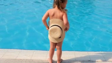 Komik küçük kız 3 yaşında havuzun ya da denizin mavi suyunun arka planında güneş şapkasının arkasına saklanıp dans ediyor. Yaz tatili konsepti, güneş kremi, seyahat. seçici odak