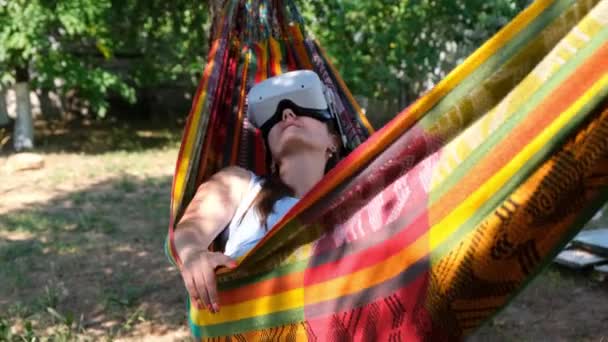 Female wearing virtual reality glasses rests in a hammock near a tree, imagining a dream come true, an invented world. The concept of getting experience using VR headset glasses of virtual reality — ストック動画