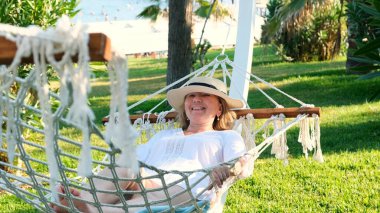 Hasır şapkalı yaşlı kadın palmiye ağaçlarının etrafındaki bir hamakta dinleniyor ve huzur ve rahatlığın tadını çıkarıyor. Yaşlılar yaşam tarzı konsepti