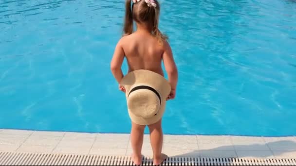 Little girl stands and covers herself with a sun hat against the backdrop of the blue water of the pool or sea. The concept of summer vacations, sunscreens, travel. selective focus on hat. — Stock Video