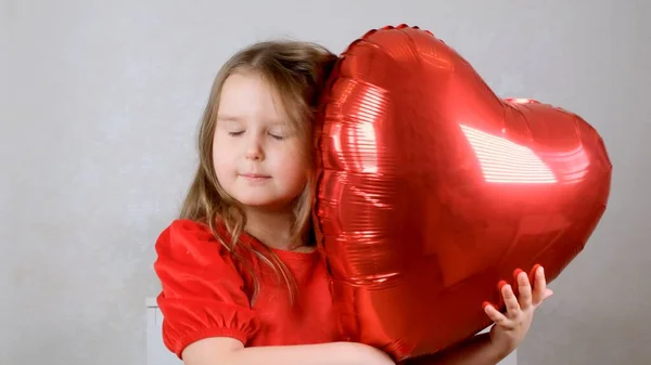 Pequena menina bonito em um vestido vermelho gentilmente abraça balões em forma de coração vermelho com as mãos. conceito dia dos namorados — Fotografia de Stock
