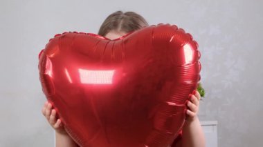 Kırmızı elbiseli küçük tatlı kız kırmızı kalp şeklinde bir balonun arkasına saklanıyor. Sevgililer Günü konsepti