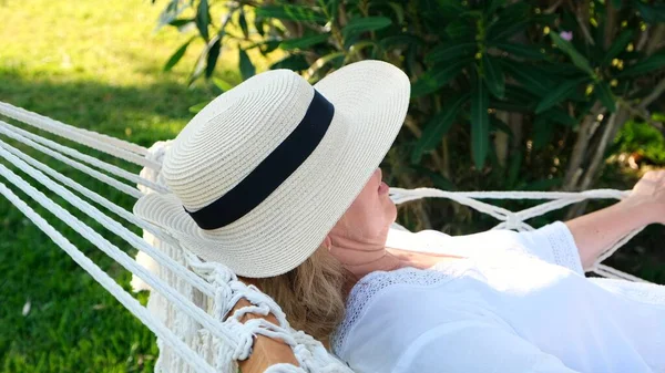 Старша жінка в солом'яному капелюсі посміхається щасливо розслабляючись на гамаку, насолоджуючись свіжим повітрям на терасі навколо пальмових дерев біля моря. Концепція способу життя старших громадян — стокове фото