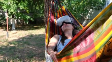Sanal gerçeklik gözlüğü takan komik kadın bir ağacın yanındaki hamakta uyduruyor. Bir hayalin gerçek olduğunu hayal ediyor. Uydurulmuş bir dünya. Sanal gözlüklerin VR kulaklığı kullanarak deneyim kazanma kavramı