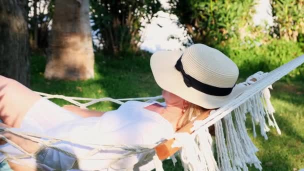 Senior woman in straw hat smiling happy relaxing on a hammock enjoying the fresh air on the terrace around palm trees near the sea. Senior citizen lifestyle concept — Wideo stockowe