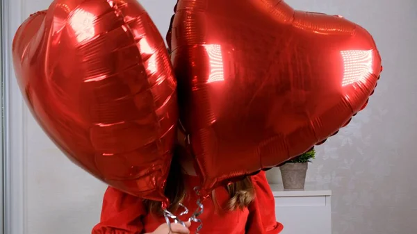 Menina engraçada em vestido vermelho escondendo atrás de três balões em forma de coração para o dia dos namorados. amor familiar. presentes para o feriado. felicidade e beijos — Fotografia de Stock